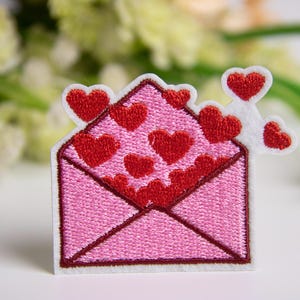 Puede incluir: Parche bordado con un diseño de sobre rosa con corazones rojos saliendo. El parche tiene un borde blanco y es adecuado para ropa o accesorios. Ideal para el Día de San Valentín o para expresar amor.