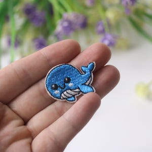 May include: A blue whale embroidered patch with white eyes and a white mouth. The whale has a white belly and a white tail fin.