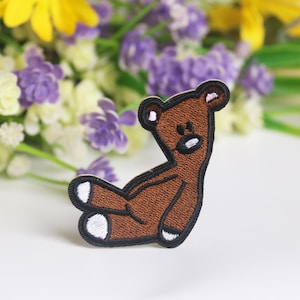 Könnte beinhalten: Ein braunes Filz-Bären-Applikationsstück mit schwarzen Nähten und rosa Nase und Ohren. Der Bär sitzt mit gekreuzten Beinen und ausgestreckten Armen.