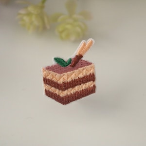 May include: An embroidered iron-on patch of a layered chocolate cake with a single green leaf and a brown stick on top.