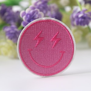 Könnte beinhalten: Rosa gestickter Smiley-Patch mit Blitzen als Augen. Der Patch hat einen weißen Rand.