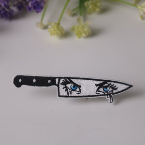 May include: White embroidered patch of a kitchen knife with two blue eyes and tears. The knife is black with a white blade.