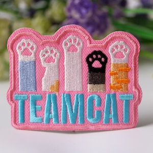 May include: Pink embroidered patch with five paw prints in different colors and the text "TEAM CAT" in blue.