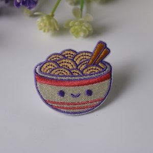 Könnte beinhalten: Eine weiße Schüssel mit rotem Rand und lila Streifen, gefüllt mit gelben Nudeln und zwei braunen Stäbchen. Die Schüssel hat ein fröhliches Gesicht.