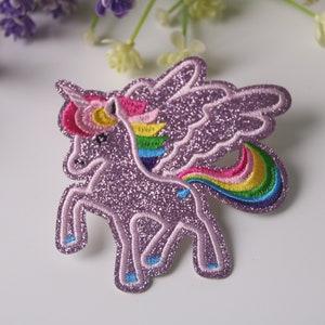 Könnte beinhalten: Ein lila glitzerndes Einhorn mit Regenbogenflügeln und -schwanz. Das Einhorn steht auf allen vieren und hat den Kopf zur Seite gedreht. Die Flügel und der Schwanz sind mit bunten Fäden bestickt.