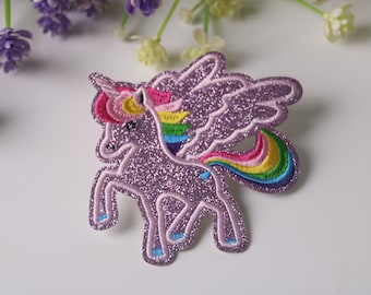 May include: A purple glitter unicorn with rainbow wings and tail. The unicorn is standing on all four legs with its head turned to the side. The wings and tail are embroidered with colorful thread.