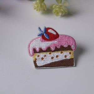 Op de afbeelding: Geborduurde strijkpatch van een roze en witte geglazuurde cake met een rode kers erop. De cake is bruin met witte glazuur en een laagje geel met bruine stippen.
