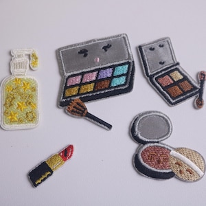 May include: Embroidered patches of makeup items, including a bottle of lotion with yellow stars, two eyeshadow palettes, a lipstick, a blush compact, and a makeup brush. The patches are in various colors.