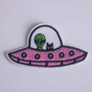 May include: A pink and white embroidered patch of a green alien and a black cat in a flying saucer.