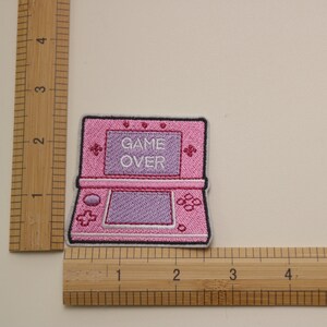 Może przedstawiać: R&oacute;żowa haftowana naszywka do prasowania przedstawiająca przenośną konsolę do gier z napisem "GAME OVER" na ekranie.