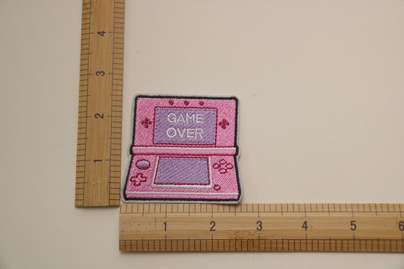 Może przedstawiać: R&oacute;żowa haftowana naszywka do prasowania przedstawiająca przenośną konsolę do gier z napisem "GAME OVER" na ekranie.