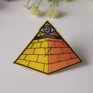 Puede incluir: Un parche bordado con una pirámide amarilla y naranja con un contorno negro y un solo ojo en la parte superior. El ojo es azul con una pupila negra y un iris blanco.