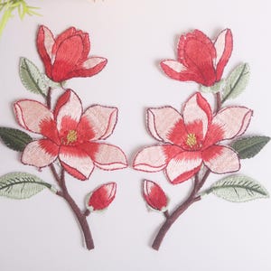 Puede incluir: Parches florales bordados con flores de magnolia rojas y rosas con hojas verdes y tallos marrones. Los parches tienen centros amarillos y están colocados sobre un fondo blanco. Parches florales decorativos.