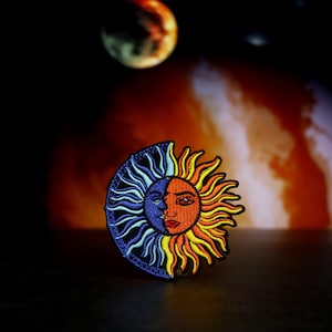 May include: A colorful embroidered patch depicting a sun and moon with faces. The sun is orange and yellow with a female face, while the moon is blue and purple with a male face.