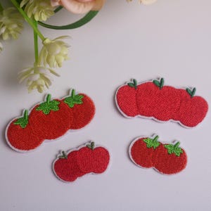 May include: Four embroidered tomato patches in various sizes. Each patch features a red tomato with green leaves and a white border. The patches are arranged on a white surface, with floral accents in the background.