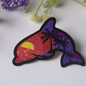 Peut inclure: Un patch brodé thermocollant représentant un dauphin violet et rouge avec un coucher de soleil et des palmiers. Le dauphin est bordé de noir.