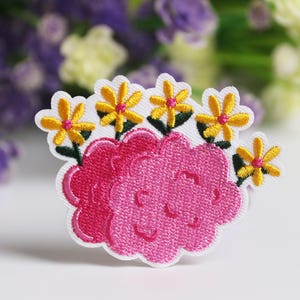 Puede incluir: Parche bordado que representa un grupo de flores rosas con centros amarillos y naranjas y tallos verdes. El parche tiene un borde blanco y está sobre un fondo borroso de flores moradas y blancas.