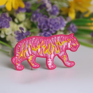 May include: A pink embroidered tiger patch with yellow flame details. The patch is in a walking pose, with a detailed design. The background features blurred flowers in yellow, purple, and white.