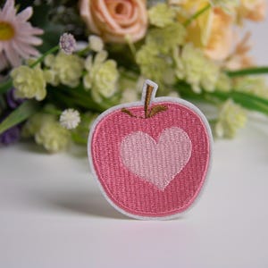Pode incluir: Um patch rosa em forma de maçã com um coração branco no centro. O patch tem uma borda branca e uma haste marrom. O fundo apresenta um buquê de flores em várias cores.