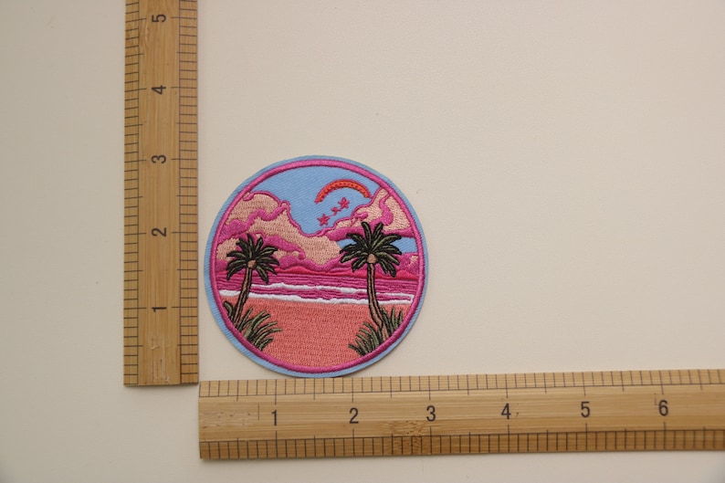 Peut inclure: Un patch rond brod&eacute; repr&eacute;sentant une sc&egrave;ne de coucher de soleil rose et bleu. Le patch pr&eacute;sente deux palmiers, une plage et un arc-en-ciel dans le ciel. Le patch mesure environ 5 cm de diam&egrave;tre.