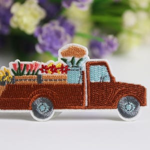 Puede incluir: Parche bordado de una camioneta marrón que transporta una variedad de flores coloridas. La camioneta tiene ventanas azules y ruedas grises. Las flores incluyen flores rojas, amarillas y azules.