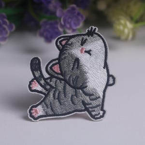 May include: An embroidered iron-on patch featuring a gray and white tabby cat in a stretching pose. The cat has closed eyes, a small pink nose, pink inner ears, and pink paw pads, with dark gray stripes and a white belly and face.
