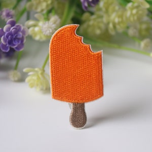 May include: Orange embroidered iron-on patch of a popsicle with a bite taken out of it.