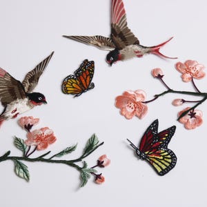 Può includere: Toppe ricamate raffiguranti uccelli, farfalle e rami fioriti. Gli uccelli hanno dettagli marroni, rossi e bianchi. Le farfalle sono arancioni, gialle e nere. I fiori sono rosa.