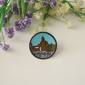 Peut inclure: Un &eacute;cusson rond brod&eacute; repr&eacute;sentant un paysage d&eacute;sertique avec des formations rocheuses brunes, des cactus et un ciel turquoise. L'&eacute;cusson a une bordure noire et est pos&eacute; sur un fond blanc avec des fleurs violettes et blanches.