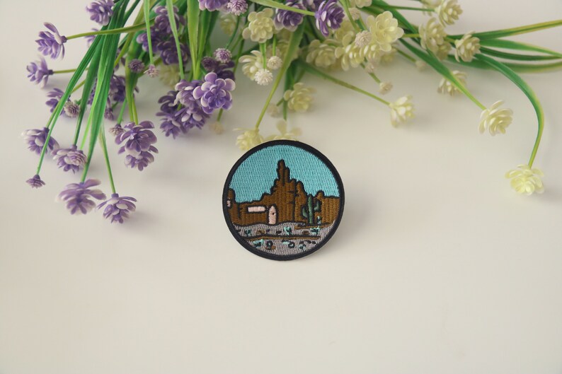 Peut inclure: Un &eacute;cusson rond brod&eacute; repr&eacute;sentant un paysage d&eacute;sertique avec des formations rocheuses brunes, des cactus et un ciel turquoise. L'&eacute;cusson a une bordure noire et est pos&eacute; sur un fond blanc avec des fleurs violettes et blanches.