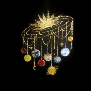 May include: An embroidered patch on a black background featuring a golden sun rising over a dark orbital ring. Multiple golden threads hang down, suspending various colorful embroidered planets, small stars, and crescent moon shapes.