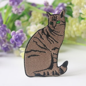 Op de afbeelding: Gestikt patch met een bruine tabby kat met groene ogen. Het patch ligt op een wit oppervlak met paarse bloemen op de achtergrond.