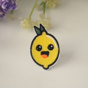 Könnte beinhalten: Ein gelber gestickter Bügelaufkleber mit einem Cartoon-Zitronenmotiv mit einem lächelnden Gesicht und schwarzen Augen. Die Zitrone hat einen grünen Stiel und Blätter.