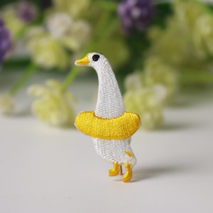 Puede incluir: Ganso blanco bordado con un salvavidas amarillo. El ganso está de pie sobre una superficie blanca.