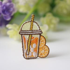 Può includere: Toppa ricamata da stirare con una bevanda ghiacciata gialla e arancione con una cannuccia e due fette d'arancia.