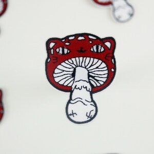 May include: Red and white embroidered iron-on patch of a mushroom with a cat face. The mushroom has white spots and a white stem.
