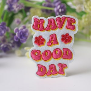 May include: A white felt patch with the phrase "HAVE A GOOD DAY" in pink bubble letters outlined in yellow. Two red embroidered flowers are also featured. The patch is a cheerful, decorative item.