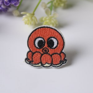 May include: An embroidered red octopus with black eyes and white pupils. The octopus has eight tentacles and is sitting on a white background.