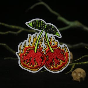 May include: Embroidered patch of two red cherries on fire with green stems and leaves. The cherries are surrounded by yellow flames. The text "Wild" is embroidered on the stems.