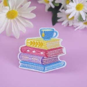 Op de afbeelding: Geborduurde patch met een stapel kleurrijke boeken met een blauwe theekop erop. De boeken zijn geel, roze, paars en blauw. Witte madeliefjes staan op de achtergrond op een roze ondergrond.