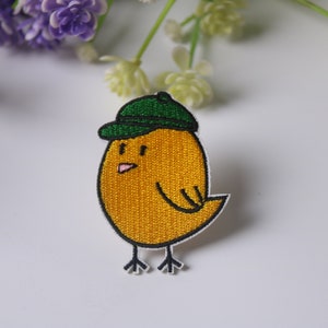 May include: A yellow embroidered chick wearing a green baseball cap. The chick has black eyes and a pink beak.
