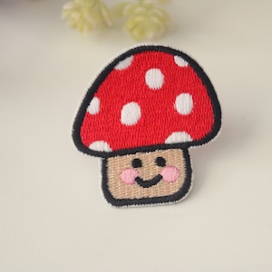 Könnte beinhalten: Gestickter Pilzaufnäher mit rotem Hut und weißen Punkten. Der Pilz hat einen beigefarbenen Stiel mit einem lächelnden Gesicht und rosa Wangen, umrandet in Schwarz. Für Kleidung oder Accessoires.