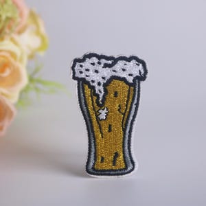 May include: An embroidered iron-on patch depicting a glass of beer with foam on top, in white and yellow.