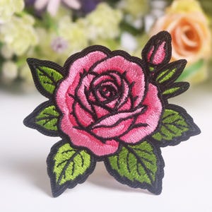 Może przedstawiać: Haftowana naszywka z różą przedstawiająca różową różę z czarnym konturem i zielonymi liśćmi. Róża ma szczegółowy wzór z pąkiem z boku. Odpowiednia do odzieży lub akcesoriów.