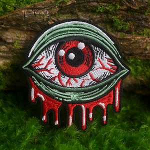 May include: An embroidered patch featuring a bloodshot eye with a bright red iris and black pupil. The eye is surrounded by a green eyelid and has red, vein-like details. Below the eye, red and white thread creates a dripping liquid effect.