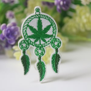 以下が含まれることがあります： 緑色のマリファナの葉が白いドリームキャッチャーに刺繍され、3本の緑色の羽根が下からぶら下がっています。