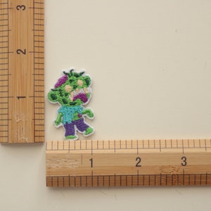 May include: Green and purple cartoon zombie embroidered iron-on patch. The zombie is wearing a blue shirt and purple pants. The patch is approximately 1 inch tall.