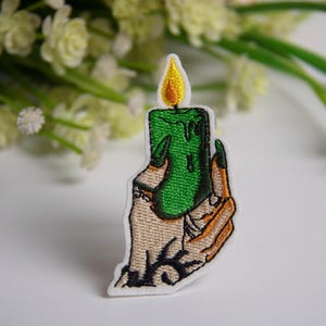 May include: Embroidered patch featuring a green candle with a yellow flame held by a tan hand with black accents. The candle has green wax dripping down. The patch has a white border and is set against a blurred floral background.