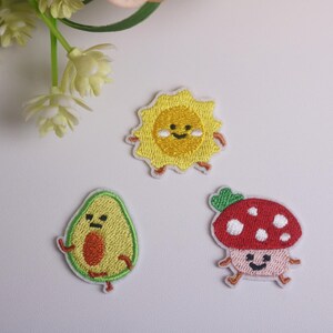 May include: Three embroidered patches: a smiling sun, an avocado, and a mushroom. The sun is yellow with a smiling face and small legs. The avocado is green with a brown pit and arms. The mushroom is red with white spots.