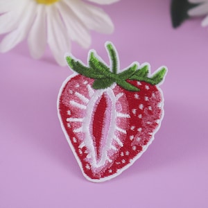 Peut inclure: Écusson brodé en forme de fraise coupée, avec des coutures rouges et blanches et des feuilles vertes. L'écusson est posé sur un fond violet clair, avec des fleurs de marguerite blanches en arrière-plan.